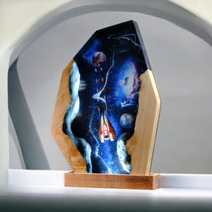 Könnte beinhalten: Ein Raumschiff fliegt durch eine Galaxie mit einem blauen, violetten und schwarzen Nebel. Das Raumschiff ist rot und weiß mit einer blauen Flammenspur. Das Kunstwerk ist in einem Holzrahmen mit einer Holzbasis gefasst.