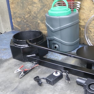 Könnte beinhalten: Eine schwarze Metall-Schmiedevorrichtung mit zylindrischem Brennergehäuse, langem Stützarm und runder Plattform. Enthält einen grauen Kunststoff-Wasserbehälter mit grünem Deckel, transparente Schläuche, ein Netzteil und ein elektrisches Gebläse.