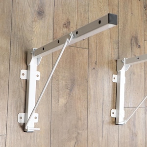 May include: Two white metal folding shelf brackets featuring a rectangular support arm with pre-drilled holes, a diagonal support strut, and a wall-mounting plate with four screw holes. The brackets have black end caps and a white finish.
