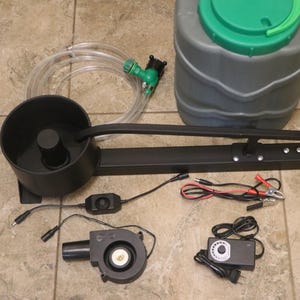 Puede incluir: Un conjunto de equipos negros y grises, que incluyen un gran recipiente redondo con tapa verde, un marco de metal negro con dos soportes cilíndricos, un soplador y varios cables y conectores. También hay una manguera transparente con una boquilla verde.