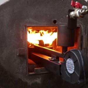 Puede incluir: Un horno de metal gris oscuro con una abertura rectangular llena de llamas de color naranja brillante. Una tubería negra y un ventilador están unidos al costado. Una válvula roja es visible en la parte superior.