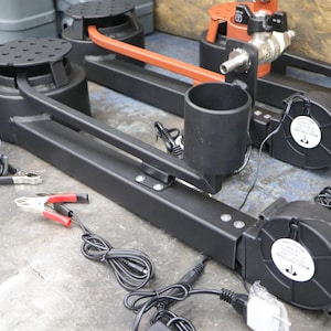 Puede incluir: Dos soportes de metal negro con un ventilador negro, un portavasos negro y una base negra. Los soportes están conectados a una fuente de alimentación con cables negros. También hay cables de puente negros y rojos en el suelo.