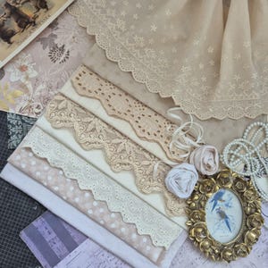 Può includere: Un assortimento di forniture artigianali in stile vintage, tra cui bordi in pizzo in varie tonalità di crema e beige, campioni di tessuto ed elementi decorativi. Una piccola cornice dorata ornata contiene un'immagine di due uccelli blu. La scena è disposta su una superficie a motivi.