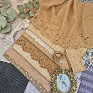 Op de afbeelding: Een verzameling beige en bruine stofstalen met verschillende kant- en oogjesontwerpen. De stoffen omvatten bloemen- en geometrische patronen, samen met een opgerolde stoffen roos en een decoratief lint. Een vintage-stijl ingelijste foto is ook aanwezig.