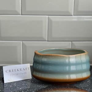 May include: A light blue ceramic bowl with a wavy rim and a brown glaze. The bowl is sitting on a black surface. The text "CREEKKRAFT" is visible on a white card in the foreground.