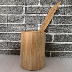 Puede incluir: Un porta utensilios de bambú natural con un utensilio de bambú a juego que descansa en su interior. El porta utensilios es cilíndrico y tiene un acabado natural liso.