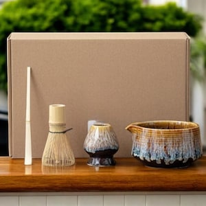 May include: A matcha tea set comprising a bamboo whisk, a ceramic bowl, a small ceramic cup, and a bamboo scoop. The set is presented on a wooden surface, with a cardboard box in the background.
