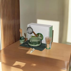 Puede incluir: Un juego de té matcha sobre una mesa de madera. El juego incluye un cuenco de cerámica verde, un batidor de bambú, un colador, una cuchara de madera y una taza de vidrio. También hay una caja blanca con un acento verde y un logotipo de té verde.