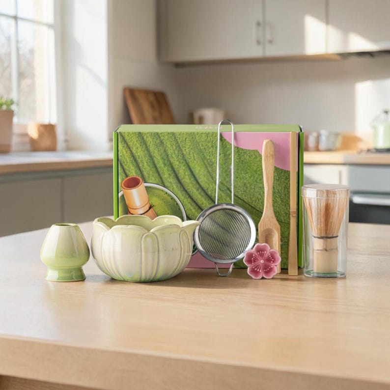 May include: A matcha tea set with a light green ceramic bowl, whisk, scoop, and sifter. The set includes a small, light green ceramic container, a pink flower-shaped ceramic piece, and a box with a green leaf design.