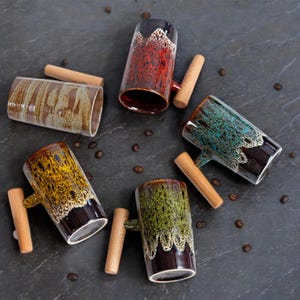 Könnte beinhalten: Fünf Keramikbecher mit Holzgriffen. Jeder Becher hat ein anderes Glasurmuster in Braun-, Grün-, Blau-, Gelb- und Rottönen. Die Becher sind in einem Kreis auf einer dunkelgrauen Oberfläche angeordnet.