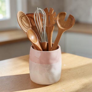 Puede incluir: Un conjunto de utensilios de cocina de madera, que incluyen cucharas y un batidor, se exhibe en un recipiente de cerámica rosa y blanco de dos tonos. El recipiente está sobre una superficie de madera clara, con una ventana al fondo.