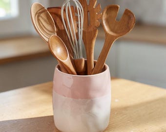 May include: A collection of wooden kitchen utensils, including spoons, a whisk, and a spatula, are displayed in a two-tone pink and white ceramic utensil holder. The holder sits on a light-colored wooden surface, with a window in the background.