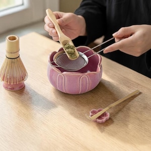 May include: A matcha tea set featuring a pink ceramic bowl, a bamboo whisk, a metal sieve, and a wooden scoop. The scoop holds green matcha powder, being sifted into the bowl. A wooden spoon rests on a pink flower-shaped holder.
