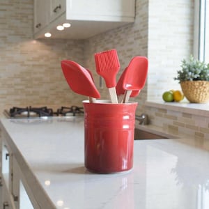 Puede incluir: Un recipiente rojo de cerámica lleno de espátulas de silicona rojas y una brocha de silicona roja. Los utensilios de cocina tienen mangos de madera. El recipiente está sobre una encimera blanca en una cocina.