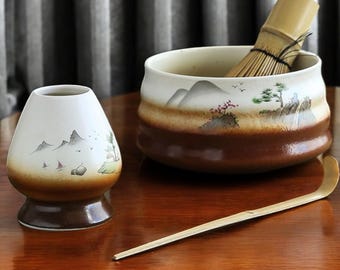 Puede incluir: Un juego de té matcha con un cuenco de cerámica blanco y marrón con un batidor de bambú, un soporte de cerámica a juego y una cuchara de bambú. El cuenco y el soporte tienen un diseño de paisaje con montañas y árboles.