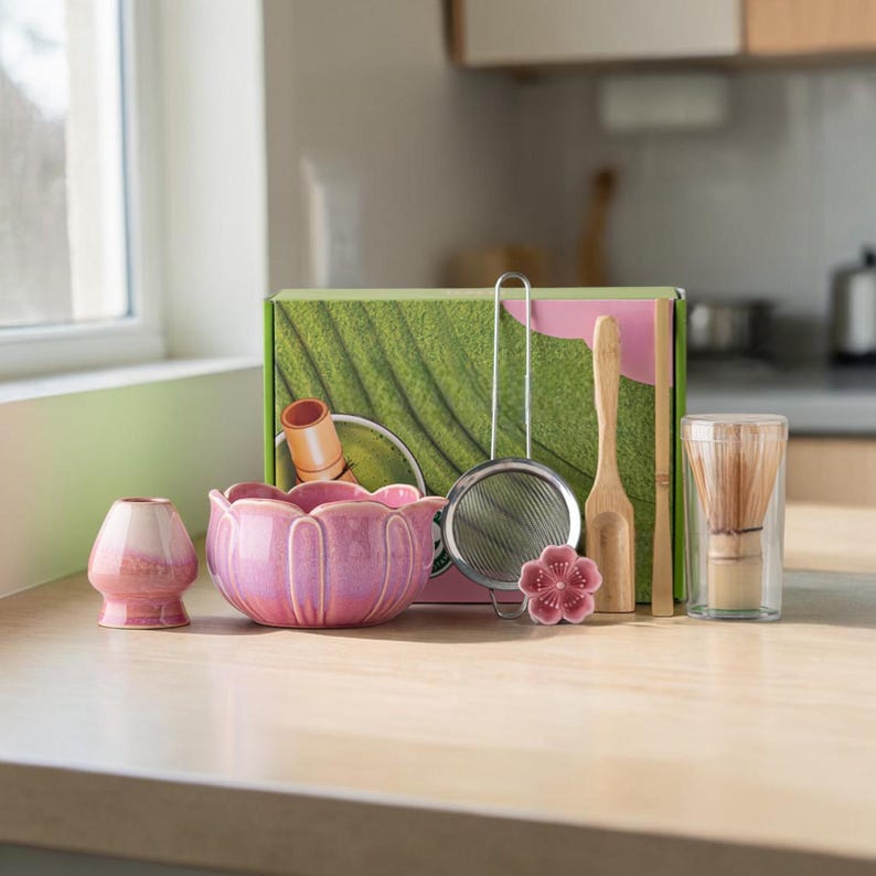 May include: A matcha tea set with a pink ceramic bowl, whisk, scoop, and sifter. The set includes a pink ceramic tea cup, a bamboo whisk, and a decorative flower. The box has a green leaf design.