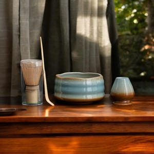 Könnte beinhalten: Ein Matcha-Teeset auf einer Holzoberfläche. Das Set beinhaltet eine blau-braune Keramikschale, einen Schneebesen in einem transparenten Behälter, einen Bambuslöffel und einen kleinen Keramikbehälter.