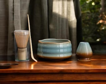 May include: A matcha tea set on a wooden surface. The set includes a blue and brown ceramic bowl, a whisk in a clear container, a bamboo scoop, and a small ceramic container.