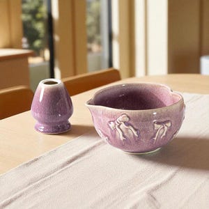 May include: A purple ceramic matcha bowl and whisk holder on a light-coloured table. The bowl has a unique shape with bow-shaped designs. The whisk holder is a small, rounded vase-like object.