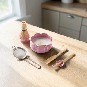 May include: A matcha tea set includes a pink ceramic bowl, a bamboo whisk, a stainless steel strainer, two wooden spoons, and a pink flower-shaped spoon rest. The set is arranged on a light-colored wooden surface.