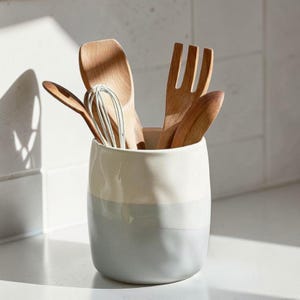 Puede incluir: Un recipiente para utensilios de cerámica de dos tonos en blanco y gris, lleno de utensilios de cocina de madera. El recipiente contiene un batidor, cucharas y un tenedor. Los utensilios de cocina son de madera clara.
