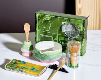 May include: A matcha tea set with a green and pink ceramic bowl, a bamboo whisk, a wooden scoop, a tea cloth, and a green box with leaf illustrations. The set includes a whisk holder and a metal sieve.