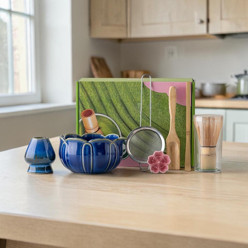 May include: A blue ceramic tea bowl and a matching blue ceramic tea cup are displayed with a tea whisk, a strainer, a scoop, and a flower-shaped decoration. A box with a green leaf design is in the background.