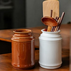 Op de afbeelding: Twee keramische bestekhouders, een bruin en een wit. De witte houder heeft houten bestek erin.