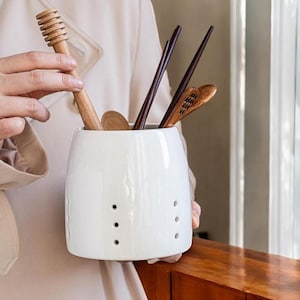 May include: White ceramic utensil holder with drainage holes, holding wooden spoons, chopsticks, and a honey dipper.