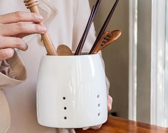 May include: White ceramic utensil holder with drainage holes, holding wooden spoons, chopsticks, and a honey dipper.