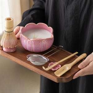 May include: A wooden tray holds a matcha tea set. The set includes a pink lotus-shaped bowl with frothy tea, a bamboo whisk, a metal strainer, a bamboo scoop, and a pink ceramic flower-shaped rest.