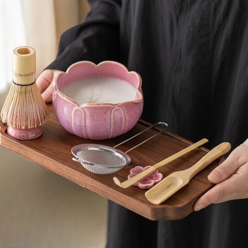 May include: A wooden tray holds a matcha tea set. The set includes a pink lotus-shaped bowl with frothy tea, a bamboo whisk, a metal strainer, a bamboo scoop, and a pink ceramic flower-shaped rest.