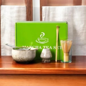 Könnte beinhalten: Ein Matcha-Teeset mit einer grünen Schachtel, einer Keramikschale, einem Schneebesen, einem Löffel und einem Chashaku. Auf der Schachtel steht "Matcha Tea Set". Das Set steht auf einer Holzoberfläche.