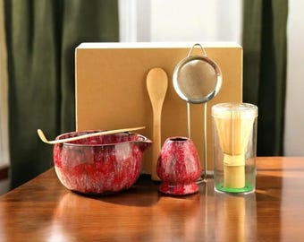 May include: A matcha tea set with a red speckled ceramic bowl, whisk, scoop, and holder. A stainless steel strainer and a wooden spoon are also included. The set is arranged on a wooden table with a cardboard box in the background.