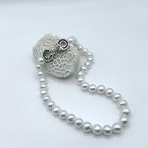 May include: A white pearl necklace with a silver clasp featuring small clear stones and two red stones. The necklace is draped over a piece of white coral, creating a classic and elegant look.