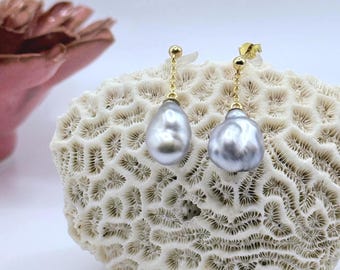 May include: A pair of elegant drop earrings featuring large, gray baroque pearls suspended from delicate gold chains. The earrings are displayed on a textured, off-white coral-like surface, with a soft pink floral element in the background.