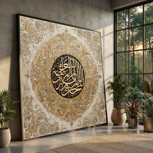 Op de afbeelding: Een groot vierkant wandkunstwerk met ingewikkelde gouden en witte filigraanpatronen rondom een centraal zwart cirkelvormig medaillon met gouden Arabische kalligrafie. Het kunstwerk is ingelijst in een dunne donkere rand.