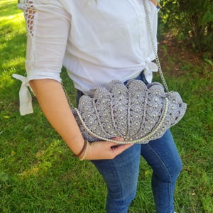 Puede incluir: Un bolso de mano de ganchillo gris con forma de concha con detalles de perlas. El bolso tiene una correa de cadena plateada.