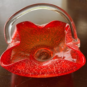 May include: A red glass basket with a clear handle. The basket has a textured surface with white speckles. The interior of the basket is a lighter shade of red. The handle is arched and attached to the sides of the basket.