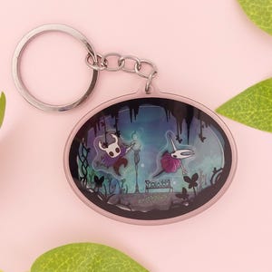 以下が含まれることがあります： ホロウナイトのキャラクター2体、ベンチ、街灯が描かれた楕円形のアクリルキーホルダー。背景はダークブルーとティールカラーで、垂れ下がるツタと蝶の黒いシルエットがデザインされています。