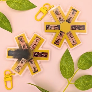 May include: Two star-shaped acrylic keychains featuring yellow borders and central illustrations of human eyes. One keychain displays a black ink-like splatter effect in the centre. Both include yellow swivel clasps for attachment.