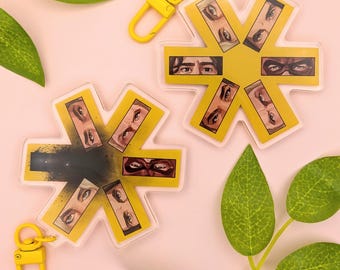 May include: Two star-shaped acrylic keychains featuring yellow borders and central illustrations of human eyes. One keychain displays a black ink-like splatter effect in the centre. Both include yellow swivel clasps for attachment.