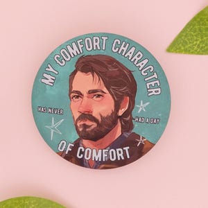 May include: A circular pinback button featuring an illustration of a man with dark hair and a beard. The teal background includes white star accents and the text: MY COMFORT CHARACTER HAS NEVER HAD A DAY OF COMFORT.