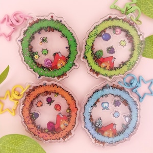 May include: Four circular acrylic shaker keychains featuring pixel art designs of a house surrounded by seasonal foliage. Each keychain has a star-shaped clasp in pink, green, yellow, or blue, and contains small loose pixel art charms inside.