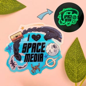 May include: A die-cut sticker featuring the text I LOVE SPACE MEDIA with a heart, a cassette tape, a xenomorph head, a space station, and a ringed planet. The design includes a glow-in-the-dark version shown in a circular inset at the top right.