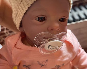 May include: A close-up of a baby doll wearing a white knitted hat and a pink shirt with a leaf design. The doll has brown eyes and is holding a clear pacifier with a white design.