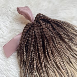 May include: Braided hair extensions in shades of brown and blonde, secured with a pink ribbon. The braids fade from dark brown at the top to a lighter blonde at the ends, creating a gradient effect. The hair extensions are styled on a white, fluffy surface.