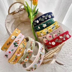 May include: A collection of embroidered headbands in various colors, including pink, yellow, green, and red. Each headband features a different floral design with white, yellow, and blue flowers. The headbands are displayed near a woven basket and tulips.