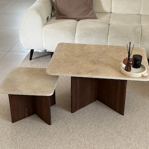 Peut inclure: Ensemble de deux tables basses. Les tables ont des plateaux en pierre beige clair et des pieds en bois brun foncé. La plus grande table est rectangulaire, et la plus petite est carrée. Elles sont sur un tapis beige.