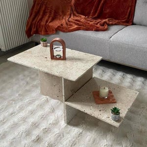 Peut inclure: Une table basse en travertin à deux niveaux avec un design géométrique. Le niveau supérieur présente un petit brûleur d'encens et une plante en pot. Le niveau inférieur a une bougie et une plante en pot.